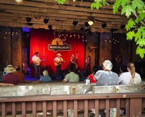 9d8262c6-050f-462d-82c2-4f5665c6868f | Branson.com | Branson.com Outdoor music stage in a rustic venue with a red curtain and a sign reading 'Silver Dollar City'; a band of four musicians plays guitars and sings while string lights hang above. Audience seated on wooden benches watches the performance.