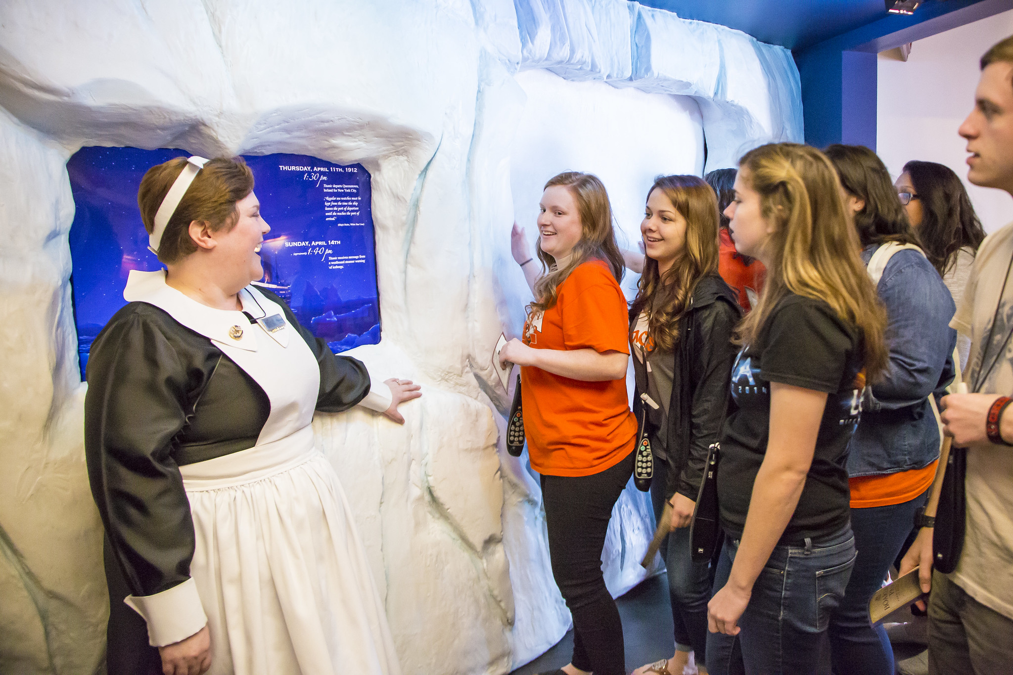 Group of visitors line up to view a large faux iceberg display as a staff member in a 19th–20th century nurse costume greets them at the left edge of the scene.