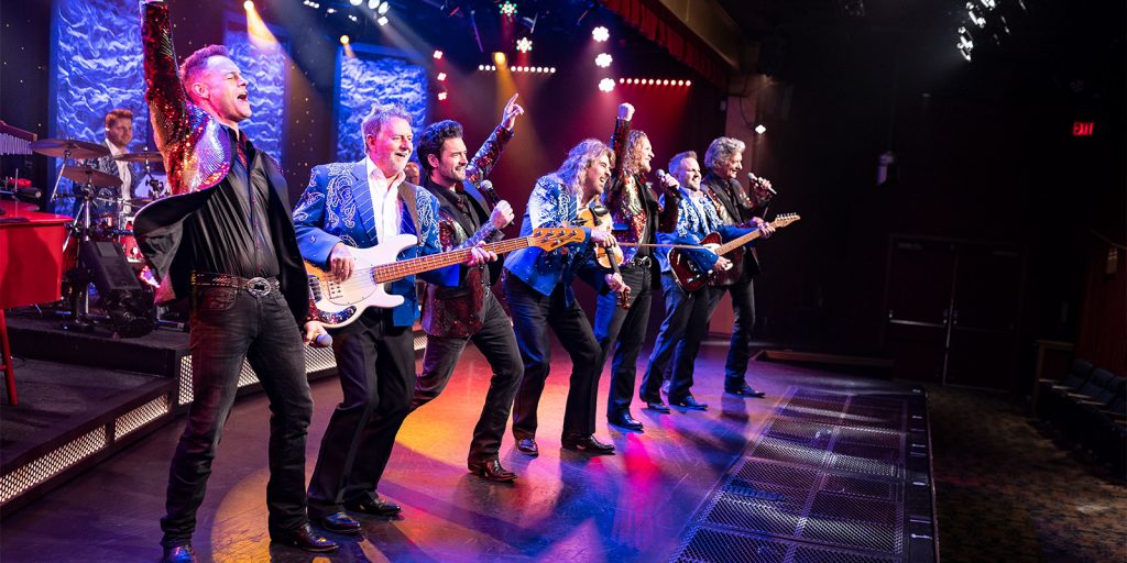 Seven-member live band on a lit stage, wearing sequined blue jackets, playing guitars and singing into microphones.