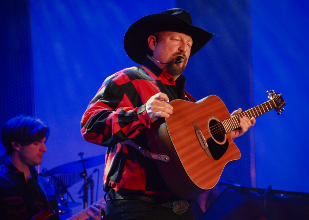 Country musician wearing a black cowboy hat and red plaid shirt plays an acoustic guitar on stage under blue lights.