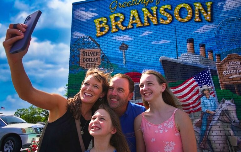 Family of four taking a cheerful selfie in front of a Branson mural with a blue sky.