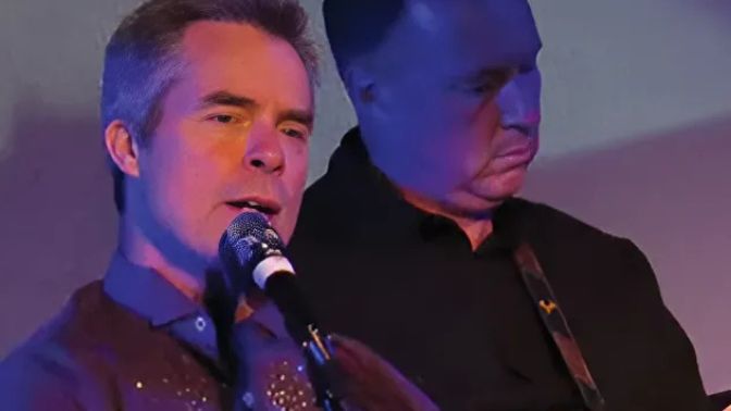 Two male performers on a dimly lit stage; one sings into a microphone while the other plays guitar, bathed in blue and purple lighting.