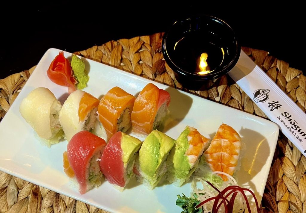 649449614_1522804976513077_1226284115453504056_n | Branson.com Assorted sushi rolls on a white plate with wasabi and pickled ginger, set on a woven placemat with a lit candle nearby.
