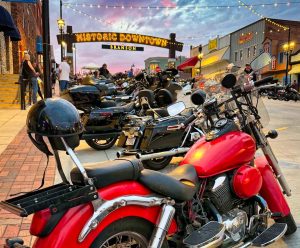6af4ac2c-252b-406b-86f7-9284c7574992 | Branson.com | Branson.com Red motorcycle in the foreground with a black helmet on the seat, parked among many bikes under string lights in Historic Downtown Branson at dusk/evening signage overhead.