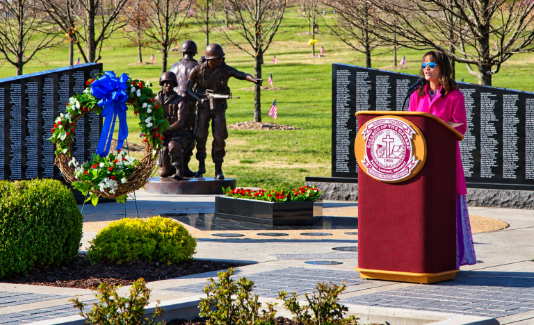 Woman in a pink dress speaks at a veterans memorial podium with a wreath and bronze soldier statues behind her.