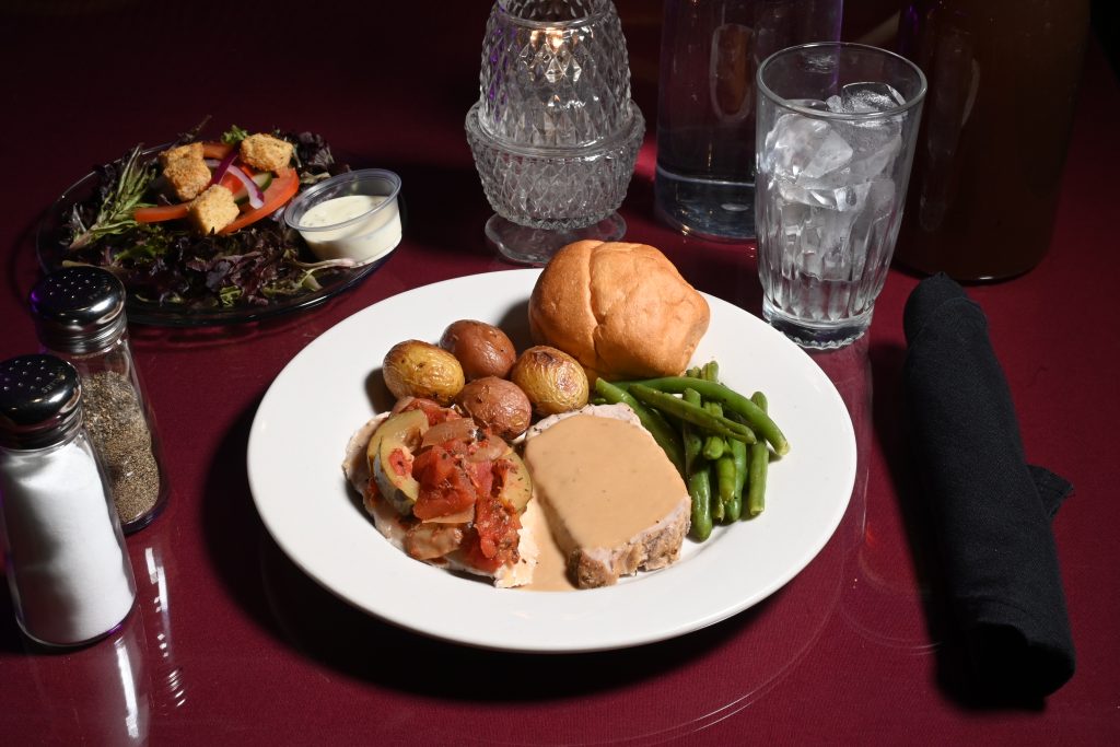 Plate of sliced turkey with gravy, roasted potatoes, and green beans on a white plate; side salad with croutons and creamy dressing on the left; burgundy tablecloth and glassware in the background.