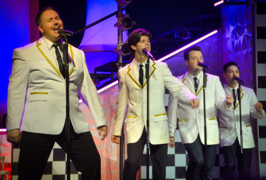 Four men in white jackets with gold trim sing into microphones on a brightly lit stage, performing together on a checkered backdrop.