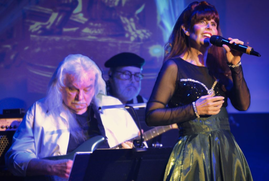Female lead singer in a black sequined dress sings into a microphone on stage, with a keyboardist in the blue-lit background nearby.