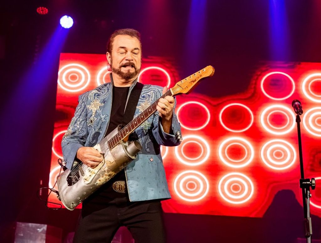Guitarist on stage in a jeweled blue jacket playing an electric guitar, with red circular LED backdrop.