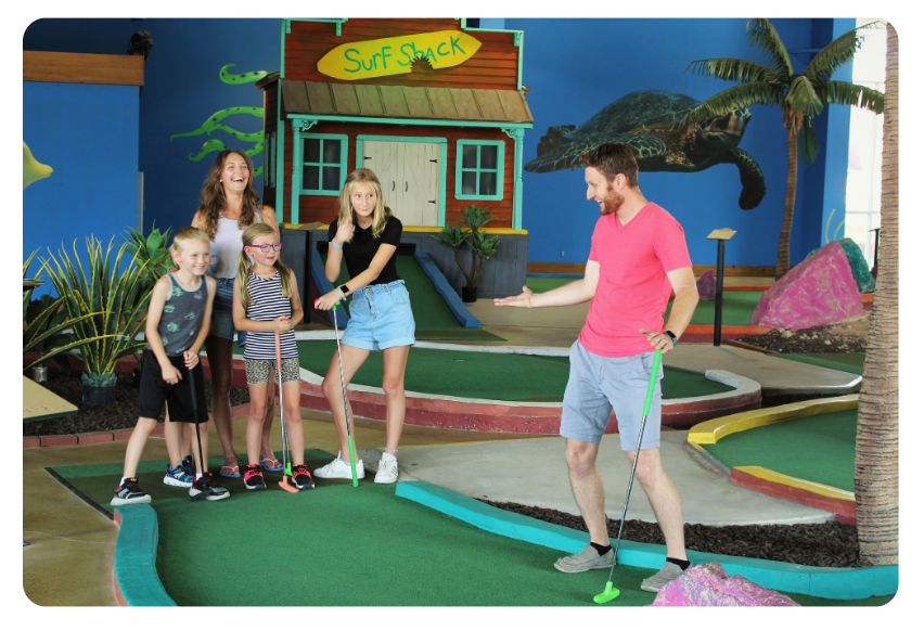 Group of children watching a man in a pink shirt give a mini-golf lesson indoors at a tropical-themed course.