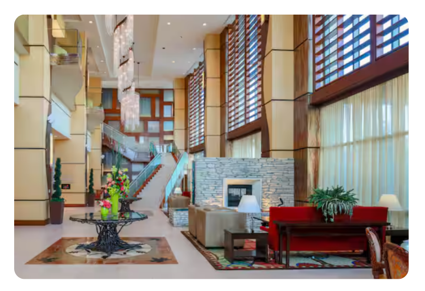 Luxurious hotel lobby with a red sofa seating area, stone fireplace, chandelier, and a glass staircase by tall windows.