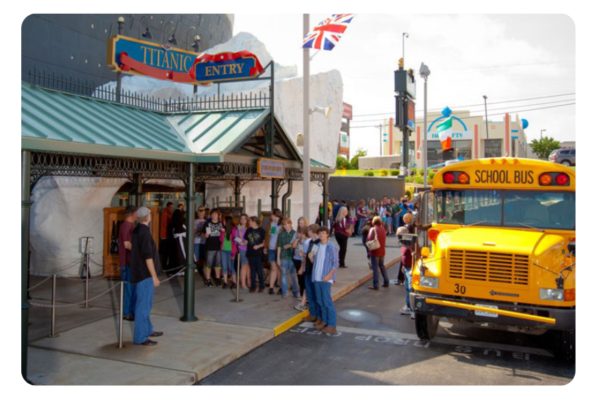 Crowd of people waiting in line outside an entrance beside a bright yellow school bus parked on the street. The scene suggests a museum or attraction area.