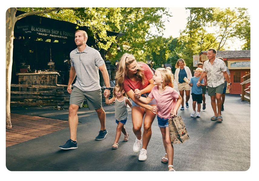 Gather Your Group 9 | Branson.com | Branson.com Family outing: parents and kids walk together along a sunny, tree-lined street, smiling and holding hands at a park area.