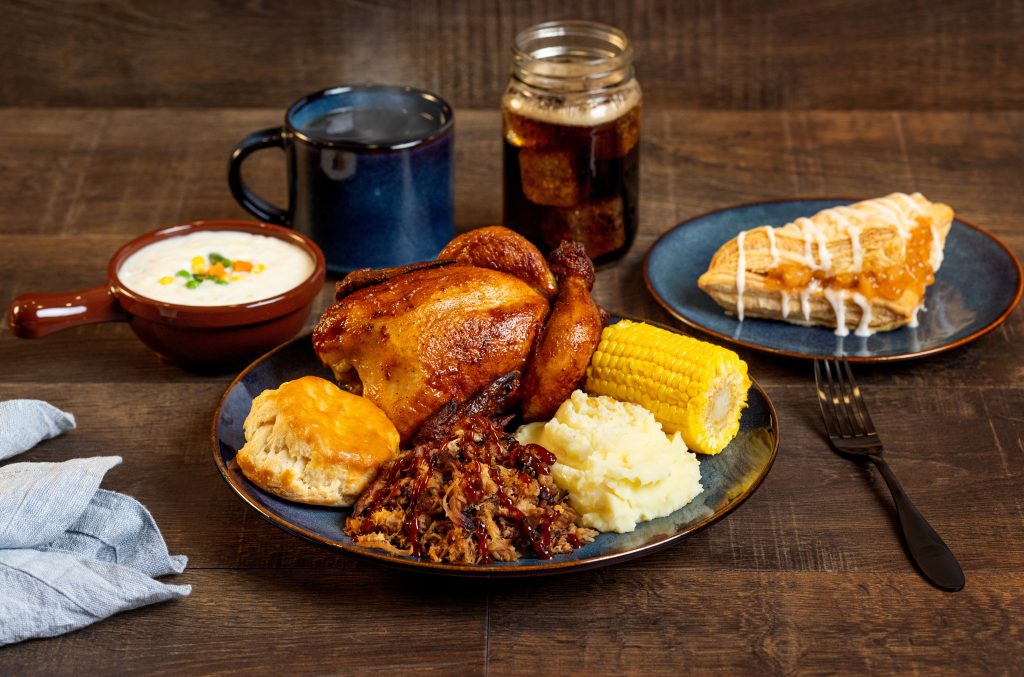 Plate of roast chicken with corn on the cob and mashed potatoes, plus a bread roll and a drizzled pastry on a blue plate; background shows a mug and a jar on a wooden table.