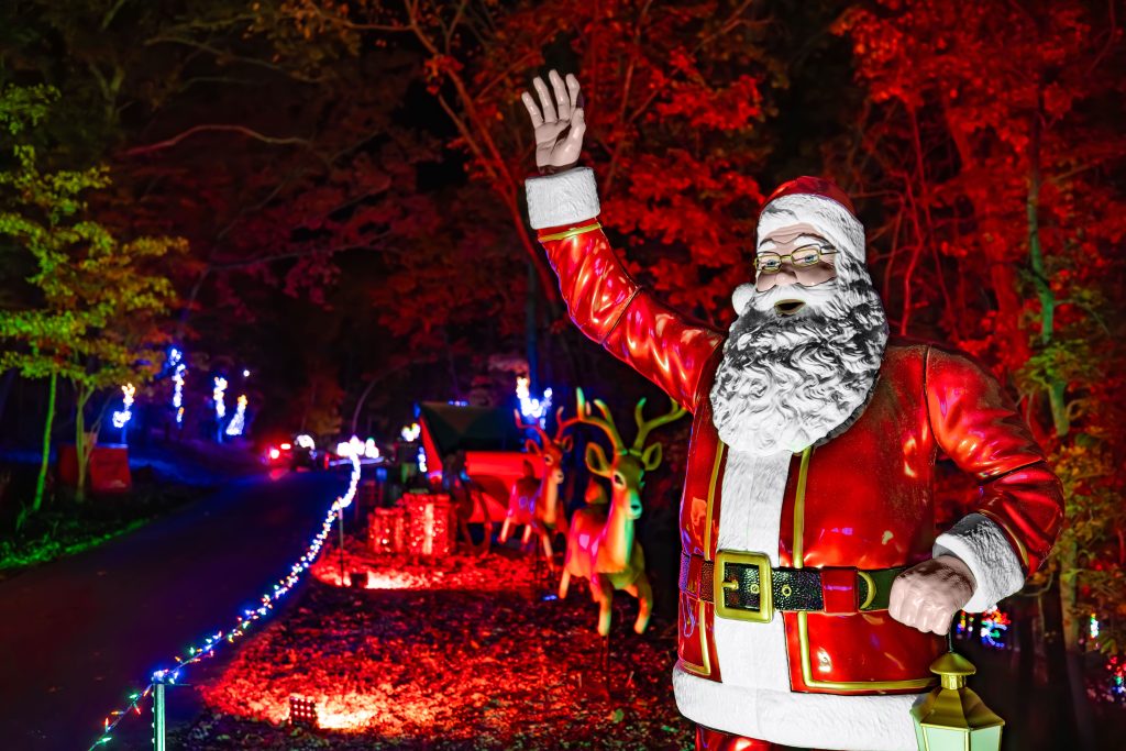 Life-size Santa Claus statue in a red suit waving at a nighttime Christmas light display.