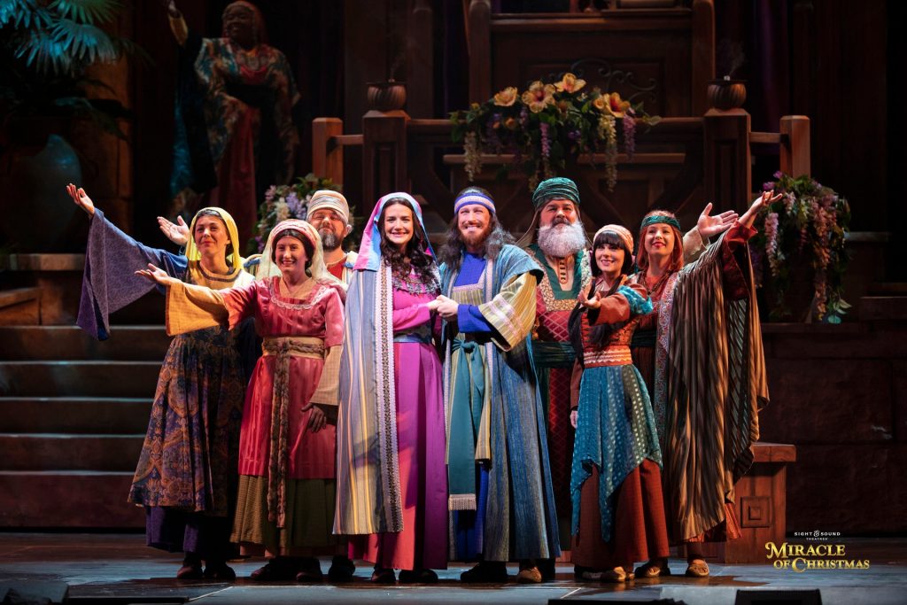 Cast of a Christmas play on stage in bright robes, arms raised in a celebratory pose.